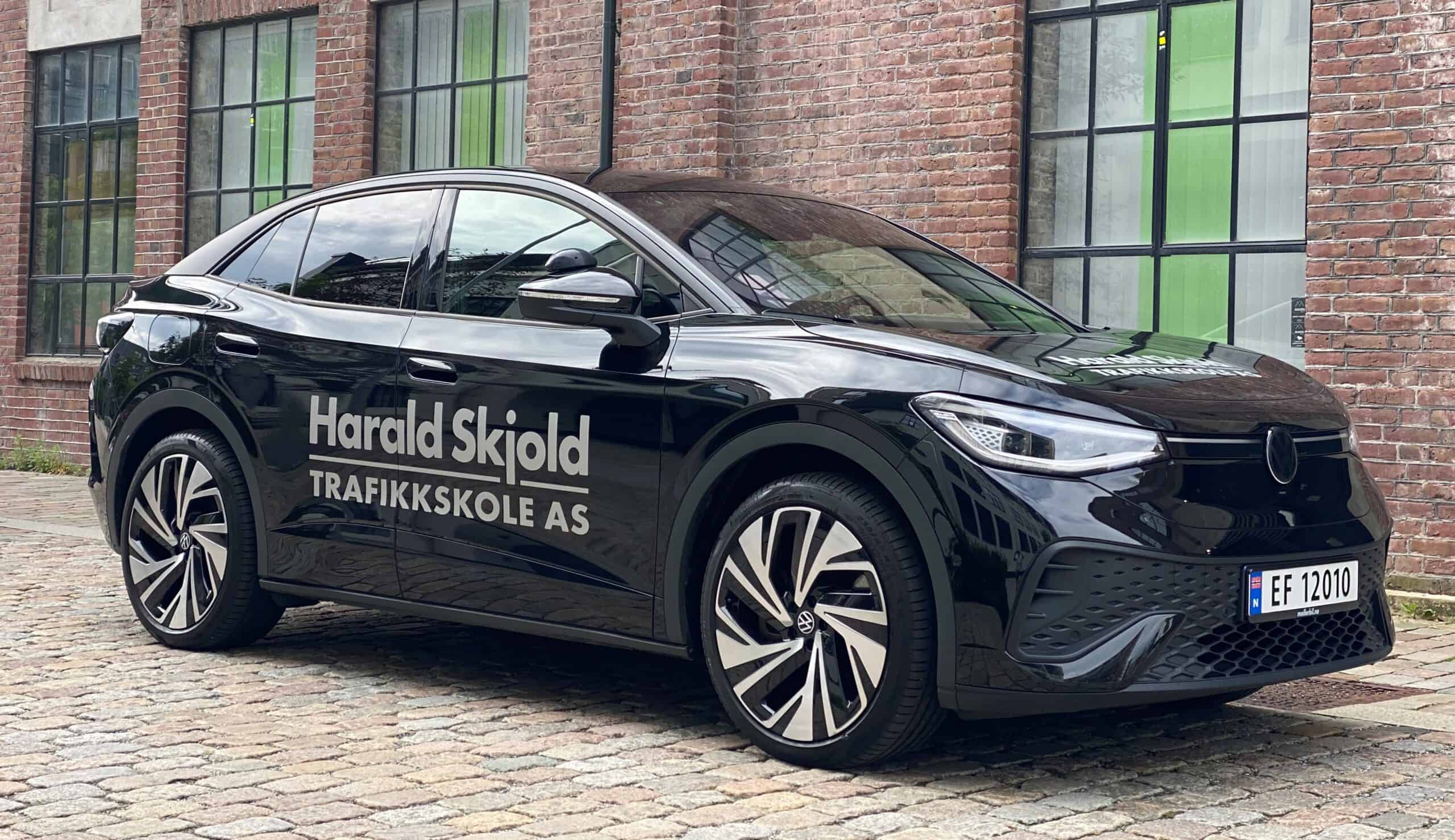 Høyde på Trafikkskolebil foran rød murvegg med store vinduer og grønne innslag, tydelig merke for Harald Skjold Trafikkskole, moderne elbil i svart med firmalogo for trafikkskole, klar for kjøreopplæring.